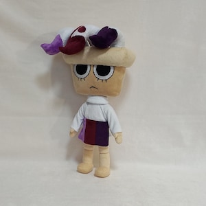May include: A plush doll with a tan head and a sad expression. The doll wears a white shirt, a skirt with purple and burgundy panels, and a hat with white, purple, and burgundy accents. The doll is approximately 12 inches tall.