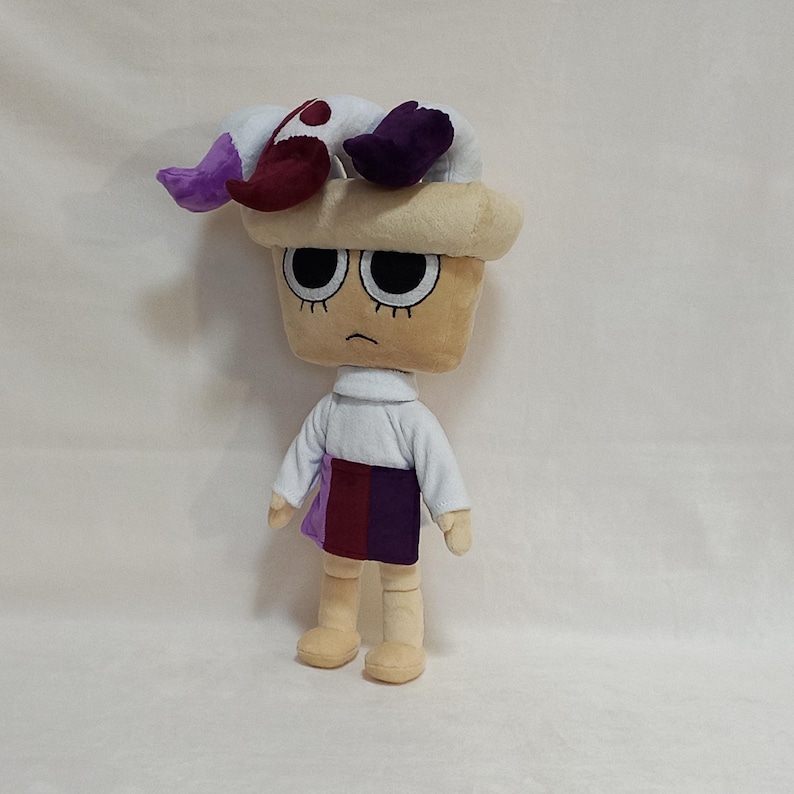 May include: A plush doll with a tan head and a sad expression. The doll wears a white shirt, a skirt with purple and burgundy panels, and a hat with white, purple, and burgundy accents. The doll is approximately 12 inches tall.