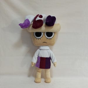 May include: A plush doll with a beige head and body, large black eyes, and a downturned mouth. The doll wears a white long-sleeved shirt, a skirt with purple and burgundy panels, and beige shoes. The doll's head has a beige hat with white, burgundy, and purple accents.