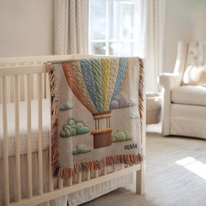 May include: A beige baby blanket with a colourful hot air balloon design, featuring a basket and clouds. The blanket is draped over a white cot. The name "Olivia" is embroidered on the blanket. The blanket has a fringed edge.