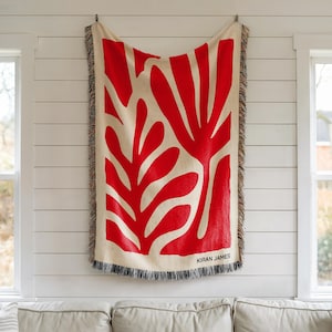 May include: A woven throw blanket with a red abstract floral design on a beige background. The blanket has a fringed edge and the name "KIRAN JAMES" is printed on the bottom right corner. The blanket is hanging on a white wall.