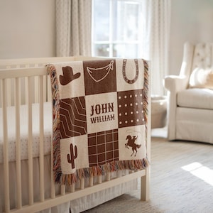 May include: A beige and brown patchwork baby blanket with cowboy-themed designs, including a hat, horseshoe, and cactus. The blanket features the name "JOHN WILLIAM" in the centre and is fringed with multiple colours. It is draped over a white cot.