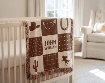 May include: A beige and brown patchwork baby blanket with cowboy-themed designs, including a hat, horseshoe, and cactus. The blanket features the name "JOHN WILLIAM" in the centre and is fringed with multiple colours. It is draped over a white cot.