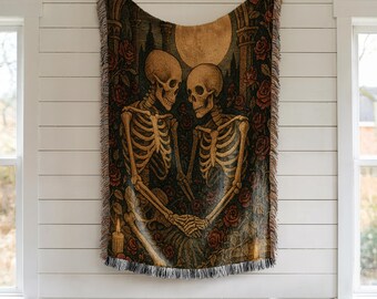 May include: A woven tapestry featuring a romantic scene with two skeletons holding hands, surrounded by roses and a full moon. The color palette includes shades of brown, black, and red. The tapestry is fringed along the bottom and is hanging on a white wall.