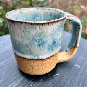 May include: A handcrafted ceramic mug with a distinctive design. The upper section has a speckled blue glaze, and the lower part is a natural tan colour. The handle and rim are also blue. Ideal for tea or coffee.