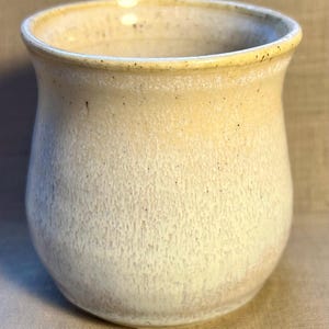 Może przedstawiać: Zestaw trzech ceramicznych kubków z nakrapianym, matowym wykończeniem. Kubki są w odcieniach kremu, jasnoszarym i ciemnoszarym. Każdy kubek ma prosty, minimalistyczny design.