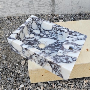 Può includere: Un lavandino rettangolare in marmo con un motivo bianco e grigio. Il lavandino ha un foro di scarico centrale e un foro più piccolo sul lato. È appoggiato su una scatola di legno.