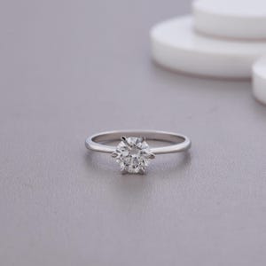 Può includere: Un anello di fidanzamento solitario con un diamante taglio rotondo incastonato in una montatura a sei griffe. L'anello ha una sottile fascia argentata lucida. Il diamante brilla su uno sfondo neutro.