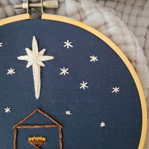 May include: Embroidered hoop art featuring a nativity scene. A large white star and smaller white stars are stitched on a dark blue background. A brown stable and manger are at the bottom. The wooden hoop is visible.