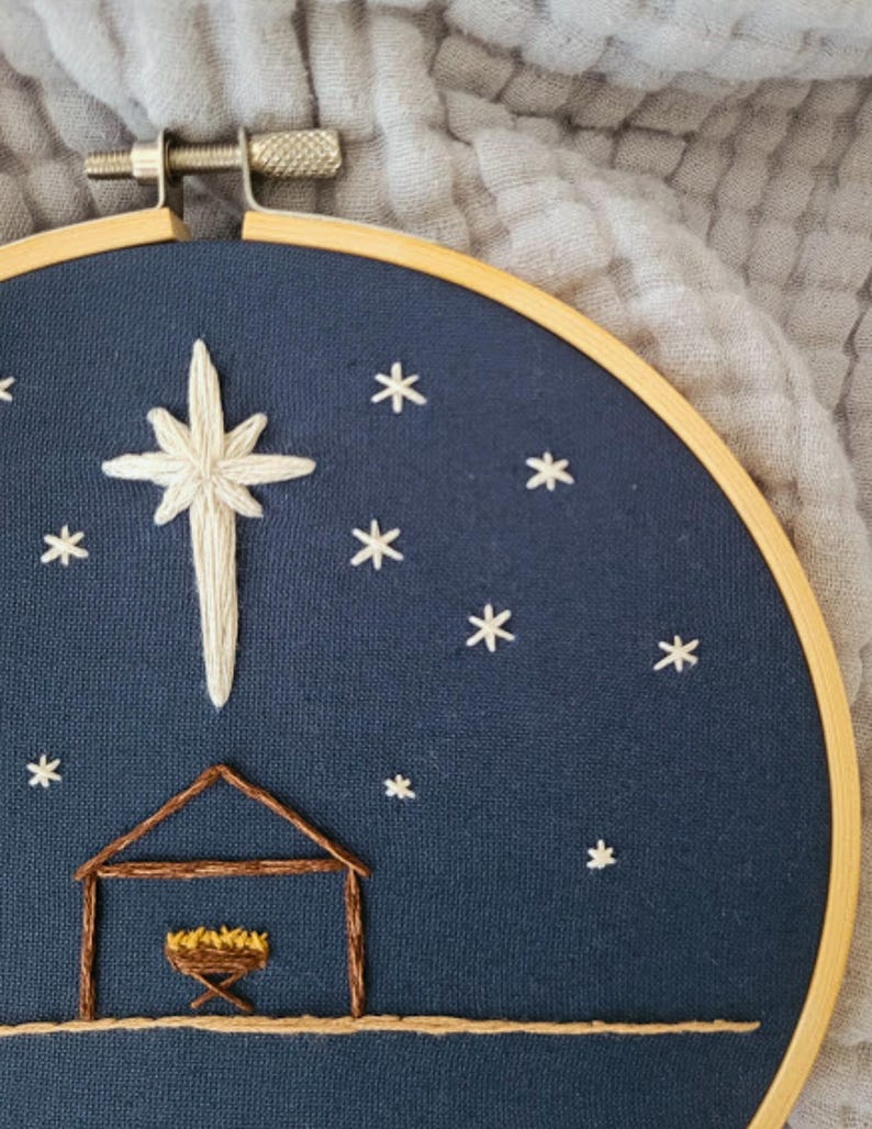 May include: Embroidered hoop art featuring a nativity scene. A large white star and smaller white stars are stitched on a dark blue background. A brown stable and manger are at the bottom. The wooden hoop is visible.