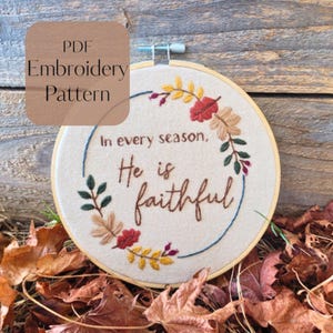 May include: A circular wooden embroidery hoop containing a beige fabric with hand-stitched text that reads In every season, He is faithful. The text is surrounded by a colorful embroidered wreath of autumn leaves in red, yellow, and brown.