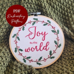 May include: A white embroidery hoop with the words "Joy to the world" in red script, surrounded by a green and red holly wreath. The words "PDF Embroidery Pattern" are in a red circle in the upper left corner.