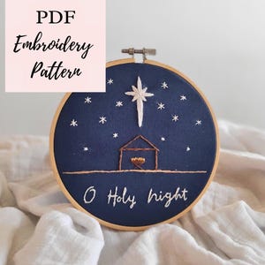 May include: A navy blue embroidery hoop with a Christmas scene. The design features a star, a stable, and the words "O Holy Night". The text "PDF Embroidery Pattern" is in a pink square.