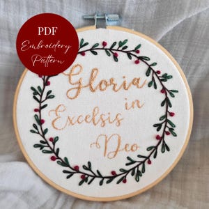 May include: Embroidered hoop art with the words "Gloria in Excelsis Deo" in gold thread. A green and red berry wreath surrounds the text. The words "PDF Embroidery Pattern" are in a red circle.