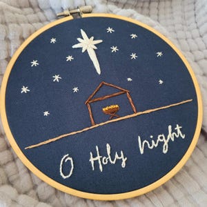 May include: Embroidered hoop art featuring a navy blue background with a white star and smaller stars. A brown stable and manger are below the star. The words "O Holy night" are stitched in white thread. The hoop is held in a wooden frame.