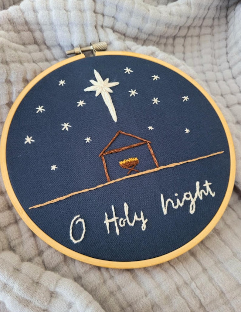 May include: Embroidered hoop art featuring a navy blue background with a white star and smaller stars. A brown stable and manger are below the star. The words "O Holy night" are stitched in white thread. The hoop is held in a wooden frame.