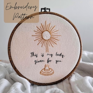 May include: An embroidery hoop featuring a religious design. The design includes a sunburst, a chalice, and the text "This is my body, given for you." The hoop is made of wood and the fabric is off-white.