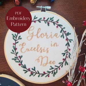 May include: A circular wooden embroidery hoop containing white fabric with the hand-stitched text Gloria in Excelsis Deo in gold thread, surrounded by a wreath of green leaves and small red berries. A red circle with white text reads PDF Embroidery Pattern.
