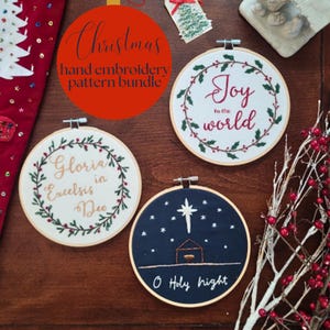 May include: Three circular wooden embroidery hoops containing fabric with stitched designs. One hoop features a green holly wreath with red berries and the text Joy to the world. Another shows a wreath with the text Gloria in Excelsis Deo. The third is dark blue with a white star, a manger, and the text O Holy night.