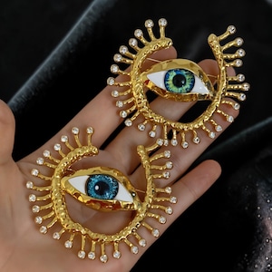 May include: Two gold-colored eye brooches. Each brooch features a detailed eye with blue and green iris, white sclera, and a gold-colored frame adorned with small, clear crystals. The brooches are held in a hand.