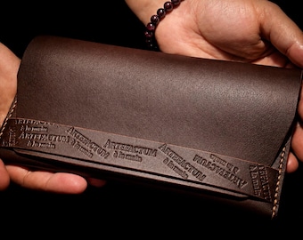 Puede incluir: Una cartera de cuero marrón oscuro con una correa horizontal grabada con el texto repetitivo ARTEFACTUM à la main. La cartera tiene costuras visibles a lo largo de los bordes laterales y un acabado mate y suave en la superficie.