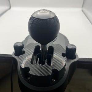 May include: A black gear shifter for a gaming setup featuring a round leather-textured knob with a silver top plate displaying a gear pattern. The base includes a carbon fibre patterned gate plate and two black cylindrical adjustment knobs.