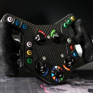 May include: A black racing wheel with a carbon fibre finish, featuring multiple coloured buttons and dials. The wheel has two black suede grips and is designed for a racing simulator setup. The wheel has a variety of buttons and dials with labels.