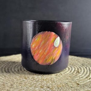 May include: A dark purple ceramic mug featuring a hand-painted planet design. The planet is a vibrant mix of orange, yellow, and red with a white accent. The mug sits on a woven mat.