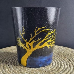 Peut inclure: Un verre peint en noir avec un motif d'arbre jaune. L'arbre est en silhouette sur un ciel étoilé. La base du verre a une zone peinte en bleu. Le verre est posé sur un tapis tissé.