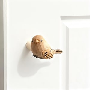Puede incluir: Un pomo de armario de madera con forma de pájaro, tallado en madera de color claro. El pájaro tiene un cuerpo redondeado, un pico pequeño y plumas de alas y cola detalladas. Está montado sobre una superficie blanca, probablemente una puerta de armario.