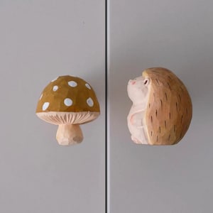 May include: Two wooden cabinet knobs: a mushroom with a brown cap and white spots, and a hedgehog with a light brown body and darker brown quills. The knobs are carved with a rustic, natural aesthetic.