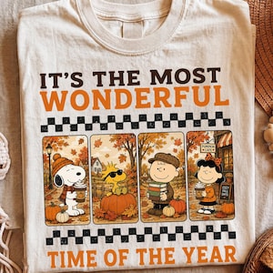 Puede incluir: Una camiseta de color crema con el texto IT'S THE MOST WONDERFUL TIME OF THE YEAR en marrón y naranja. El diseño incluye cuatro paneles que muestran a Snoopy, Woodstock, Charlie Brown y Lucy en un entorno otoñal con calabazas.