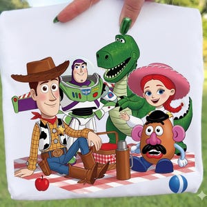 K&ouml;nnte beinhalten: Ein wei&szlig;es T-Shirt mit einem farbenfrohen Grafikdruck der Toy Story-Figuren Woody, Buzz Lightyear, Rex, Jessie und Mr. Potato Head bei einem Picknick auf einer rot-wei&szlig; karierten Decke mit einem roten Apfel und einem blauen Ball.