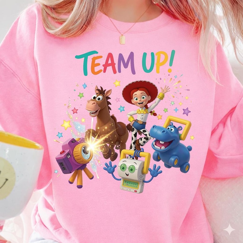 K&ouml;nnte beinhalten: Ein hellrosa Sweatshirt mit dem Schriftzug TEAM UP! in bunten Buchstaben &uuml;ber einer Grafik von Toy Story-Figuren, darunter Jessie, Bullseye, eine lila Kamera, ein blaues Nilpferd-Spielzeug und ein wei&szlig;er Roboter, umgeben von bunten Sternen.