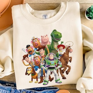 K&ouml;nnte beinhalten: Ein cremefarbenes Sweatshirt mit Rundhalsausschnitt und einem farbenfrohen Grafikdruck der Toy Story-Figuren, darunter Woody, Buzz Lightyear, Jessie, Rex, Slinky Dog, Mr. und Mrs. Potato Head, Bullseye und Hamm, die zusammenstehen.