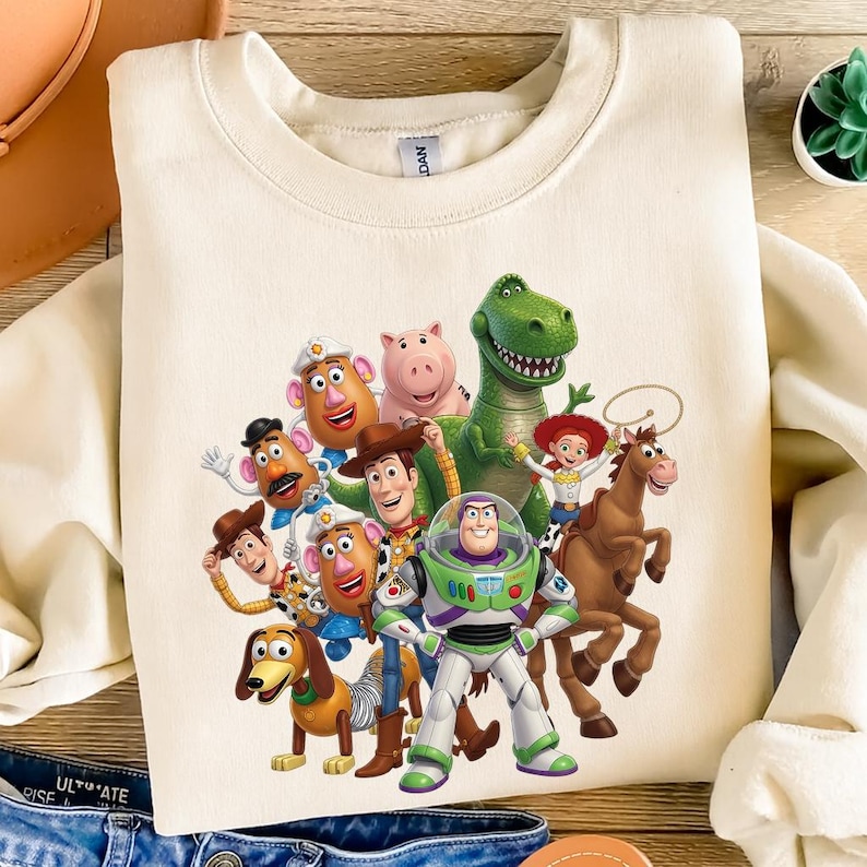 K&ouml;nnte beinhalten: Ein cremefarbenes Sweatshirt mit Rundhalsausschnitt und einem farbenfrohen Grafikdruck der Toy Story-Figuren, darunter Woody, Buzz Lightyear, Jessie, Rex, Slinky Dog, Mr. und Mrs. Potato Head, Bullseye und Hamm, die zusammenstehen.