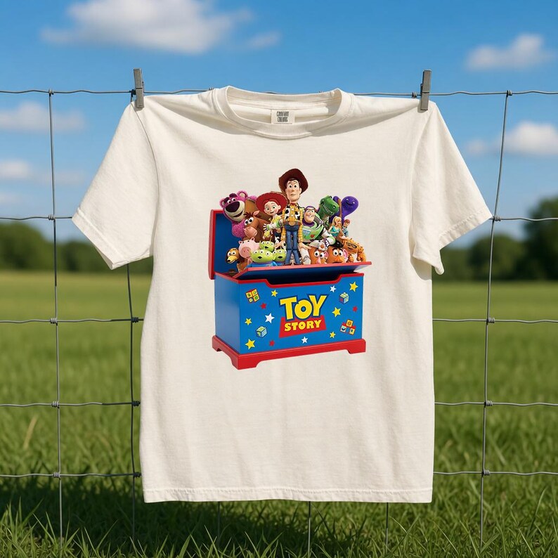 K&ouml;nnte beinhalten: Ein cremefarbenes Kurzarm-T-Shirt mit einem Grafikdruck einer blauen Spielzeugkiste mit rotem Rand. Die Kiste ist mit Toy Story beschriftet und mit verschiedenen bunten Zeichentrickfiguren gef&uuml;llt, darunter ein Cowboy und ein Weltraum-Ranger.