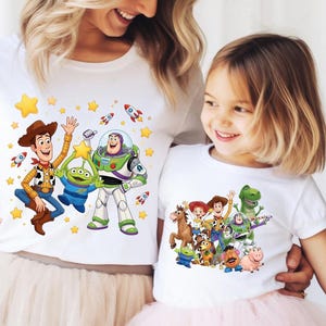 K&ouml;nnte beinhalten: Zwei wei&szlig;e kurz&auml;rmelige T-Shirts mit farbenfrohen Grafikdrucken von Toy Story-Figuren. Das gr&ouml;&szlig;ere T-Shirt zeigt Woody, Buzz Lightyear und einen Alien mit gelben Sternen und Raketen. Das kleinere T-Shirt zeigt eine Gruppe von Toy Story-Figuren.