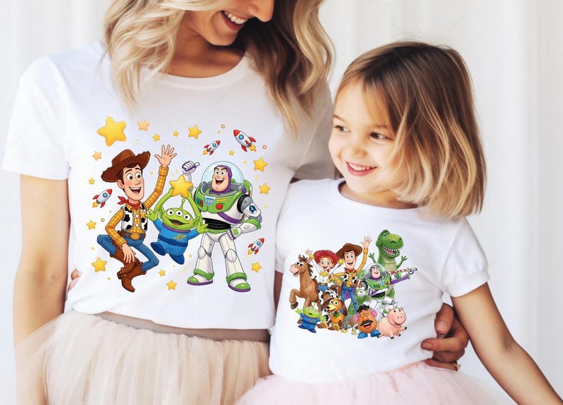 K&ouml;nnte beinhalten: Zwei wei&szlig;e kurz&auml;rmelige T-Shirts mit farbenfrohen Grafikdrucken von Toy Story-Figuren. Das gr&ouml;&szlig;ere T-Shirt zeigt Woody, Buzz Lightyear und einen Alien mit gelben Sternen und Raketen. Das kleinere T-Shirt zeigt eine Gruppe von Toy Story-Figuren.