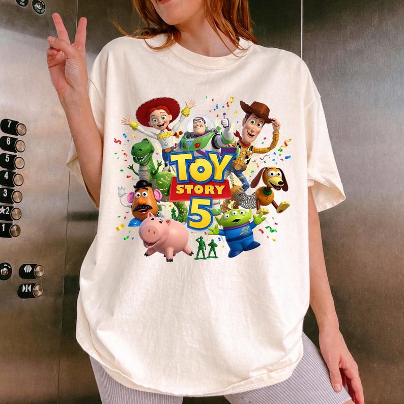 K&ouml;nnte beinhalten: Ein cremefarbenes Kurzarm-T-Shirt mit einem bunten Grafikdruck der Toy Story-Figuren, darunter Woody, Buzz Lightyear, Jessie, Rex, Slinky Dog, Mr. Potato Head, Hamm und Aliens, sowie dem Schriftzug Toy Story 5 und bunten Konfetti-Akzenten.