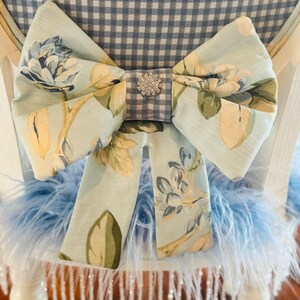 May include: A light blue floral bow with a rhinestone centre decorates a white chair. The bow is made of fabric with a floral pattern in shades of blue, yellow, and green. The chair back has a blue and white gingham pattern.