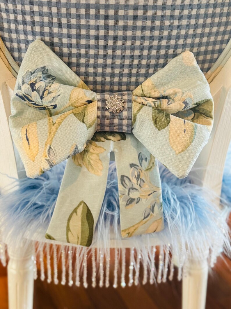 May include: A light blue floral bow with a rhinestone centre decorates a white chair. The bow is made of fabric with a floral pattern in shades of blue, yellow, and green. The chair back has a blue and white gingham pattern.