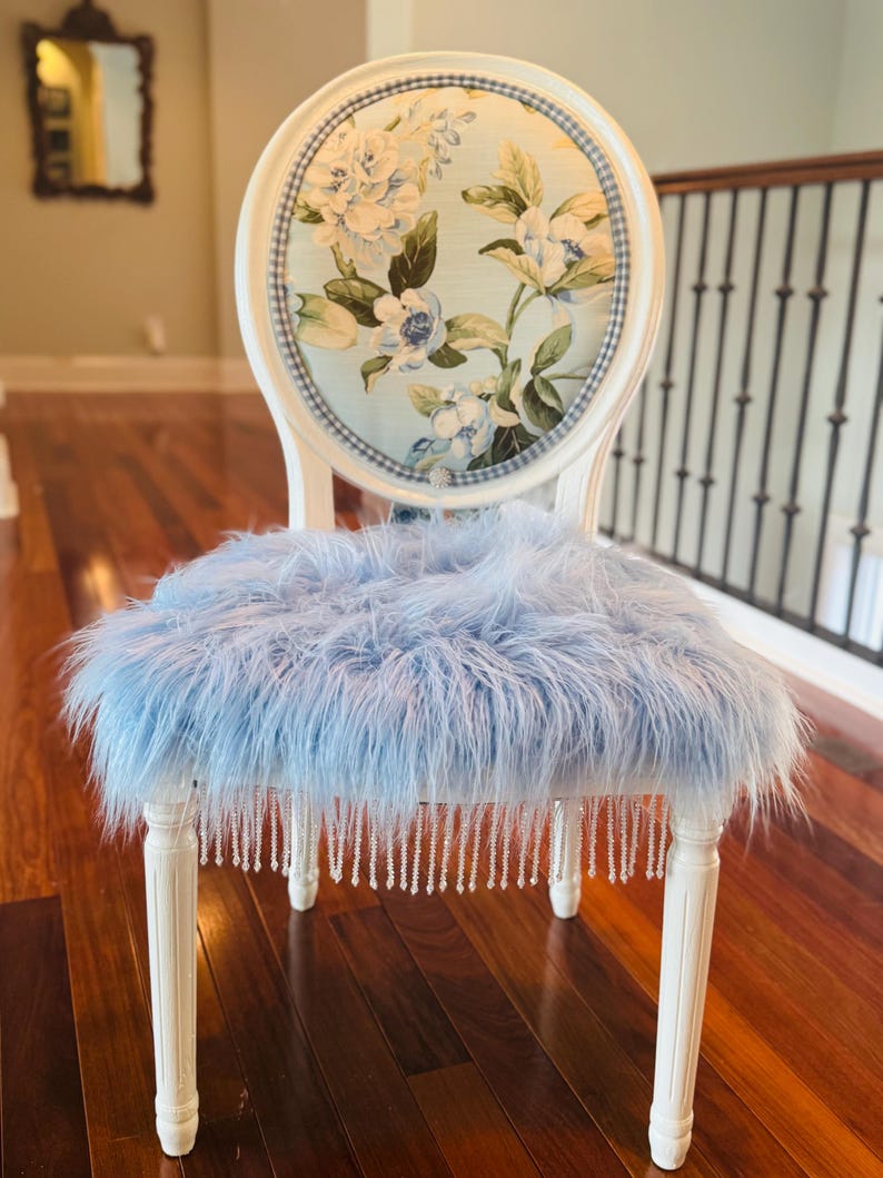 May include: An elegant white chair with a floral patterned back in blue and white. The seat is covered in light blue faux fur, and the legs are decorated with hanging crystal beads. The chair is a decorative item, suitable for a bedroom or living room.