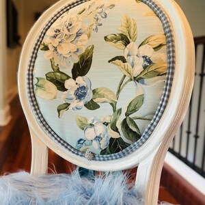 May include: A white-painted chair with a floral-patterned back. The oval back features a light blue fabric with white and blue flowers and green leaves. A blue and white checkered trim surrounds the floral fabric. The chair's seat is covered in light blue feathers.