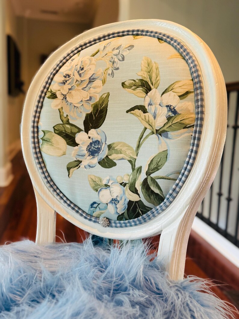 May include: A white-painted chair with a floral-patterned back. The oval back features a light blue fabric with white and blue flowers and green leaves. A blue and white checkered trim surrounds the floral fabric. The chair's seat is covered in light blue feathers.