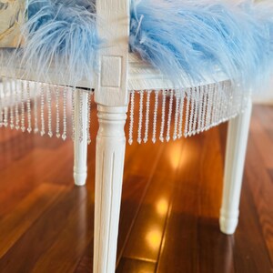 May include: A white painted chair with a light blue feather cushion and a beaded fringe. The chair legs are also white. The chair is a decorative piece, possibly for a bedroom or boudoir.