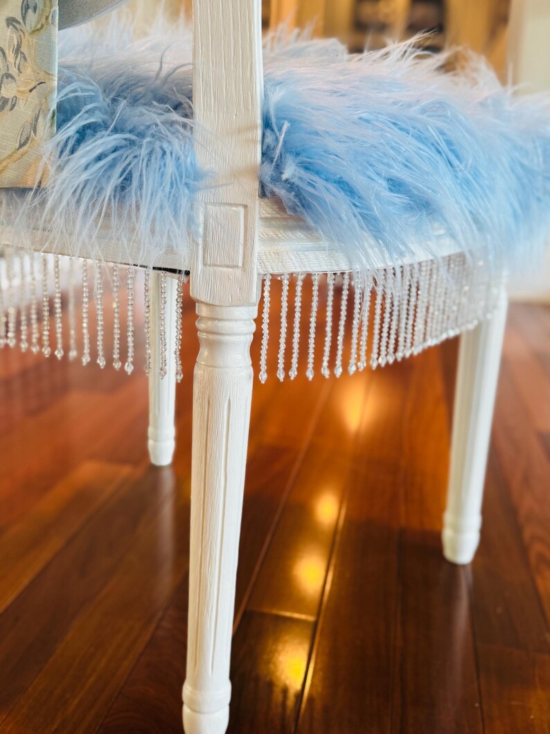 May include: A white painted chair with a light blue feather cushion and a beaded fringe. The chair legs are also white. The chair is a decorative piece, possibly for a bedroom or boudoir.