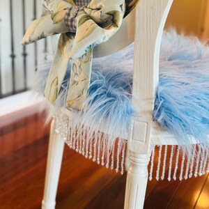 May include: A white painted chair with a blue feather seat and a floral bow. The chair back has a gingham pattern. The seat trim has hanging crystal beads. The bow has a rhinestone centre.