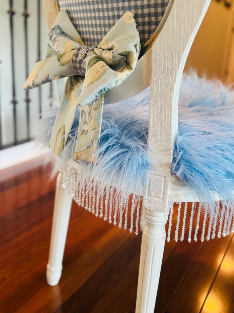 May include: A white painted chair with a blue feather seat and a floral bow. The chair back has a gingham pattern. The seat trim has hanging crystal beads. The bow has a rhinestone centre.