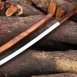 Op de afbeelding: Een Japanse katana-zwaard met een houten schede en handvat. Het lemmet is zilverkleurig en de schede en het handvat zijn diepbruin. Het zwaard ligt op een stapel houtblokken.